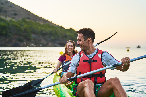 Happy man and woman talking while kayaking on river. Couple is spending leisure time during sunset. They are enjoying vacations.