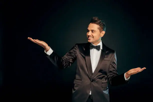Professional showman wearing suit standing isolated on black background showing copy space aside smiling joyful