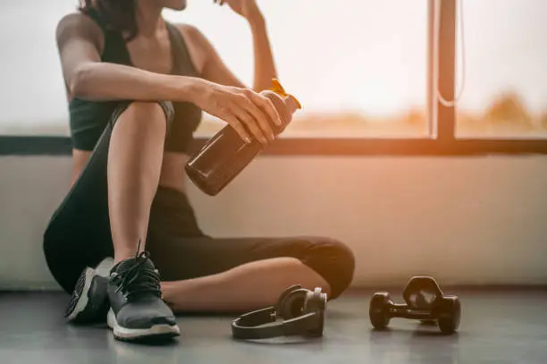Fitness woman Relaxing after exercise with a whey protein and dumbbell placed beside the gym.Relaxing after training.beautiful young woman looking away while sitting  at gym.