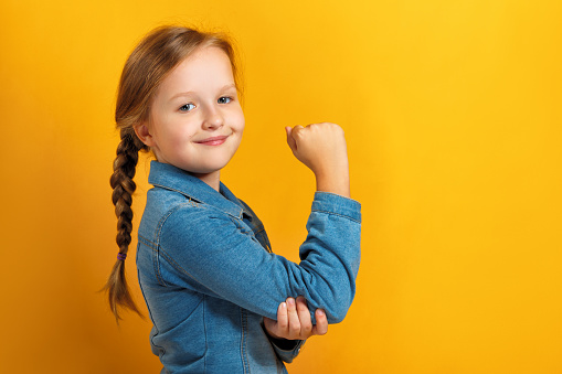 Closeup portrait of a little girl on a yellow background. Children's hand shows biceps. Girl power concept