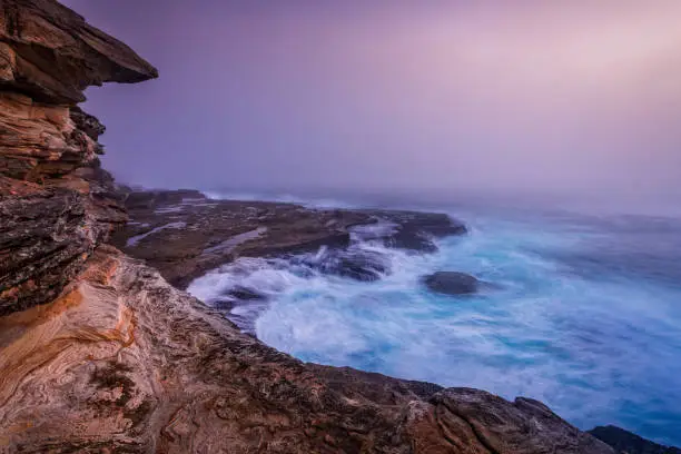 Tumultuous waves crash and cascade off the rocks under the cliffs near Sydney as a dense thick fog cut visibility,  hiding the urban landscape and buildings in the distance.