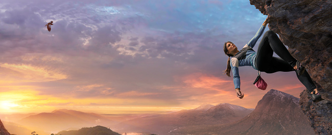 A female free climber wearing climbing shoes, leggings, top and chalk bag scaling an inclined rock face at high altitude under a beautiful dawn sky.  The climber has two feet and one outstretched arm in contact with the rock face and is leaning back with other arm relaxed.