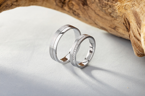 Pair of silver wedding rings with diamonds on gray background with wood. White gold wedding rings band with matte surface and gemstones