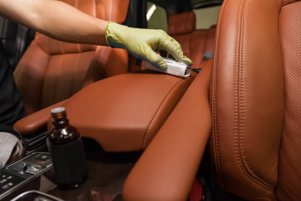 Applying a nano-ceramic coating for interior leather on the car's seat brown upholstery by a worker in yellow gloves with a sponge and bottle of chemical composition. Auto service industry. aplicación de un recubrimiento nanocerámico para cuero interior en la tapicería marrón del asiento del coche por un trabajador con guantes amarillos con una esponja y una botella de composición química. industria de servicios automotriz.
