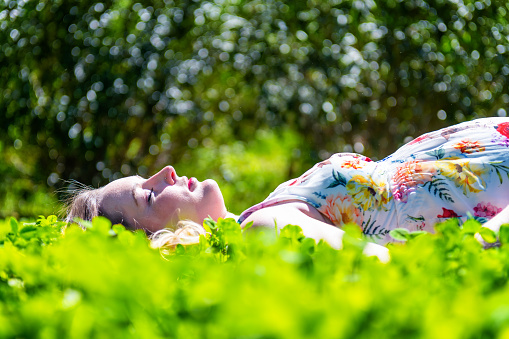 Beautiful pregnant woman lying down in grass and wearing floral dress. Young mother is surrounded by idyllic nature in summer. Future mother relaxing with eyes closed, napping, smiling and touching her belly. Beauty in life and lots of love for baby on the way.