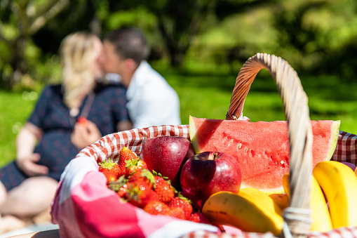 Picnic basket with choice of organic fruits arranged in park. Fresh apples, strawberries, bananas and watermelon inside of picnic basket. Lovely defocused couple kissing in the background.
