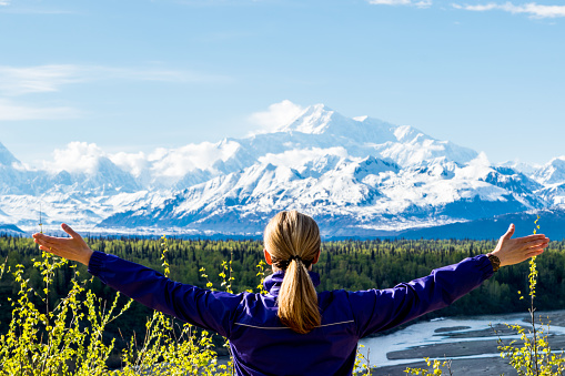 Woman with arms outstretched looking at Mount Denali in Alaska by a sunny day.