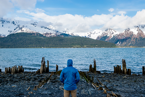 Man looking at a rotten wharf by the Resurrection Bay and the city of Seward, Alaska, with snowcapped mountains in background.