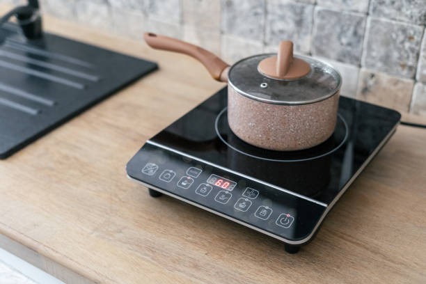 Kitchenware pan at small electric stove with timer on control panel. Modern kitchen with wooden surface table and marble wall tile at blurred background Kitchenware pan at small electric stove with timer on control panel. Modern kitchen with wooden surface table and marble wall tile at blurred background glass ceramic stove top stock pictures, royalty-free photos & images