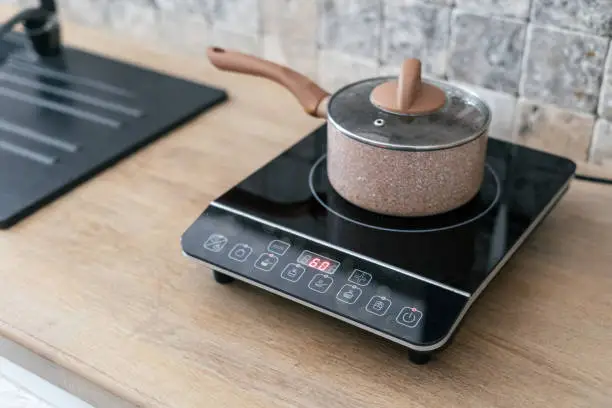 Photo of Kitchenware pan at small electric stove with timer on control panel. Modern kitchen with wooden surface table and marble wall tile at blurred background