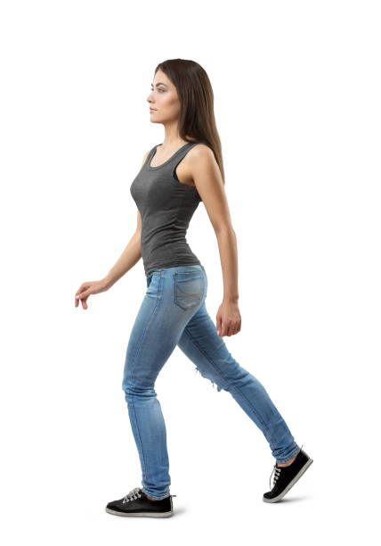 Side view of young woman in gray sleeveless top and blue jeans, with long dark hazelnut hair, walking forward on white background. Side view of young woman in gray sleeveless top and blue jeans, with long dark hazelnut hair, walking forward on white background. Modeling business. Catwalk show. Feel confident and attractive. catwalk stage stock pictures, royalty-free photos & images