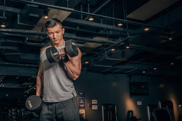 Fit Chinese guy practicing with dumbbells in gym One Chinese man, training with dumbbell alone in dark gym. weightlifting stock pictures, royalty-free photos & images