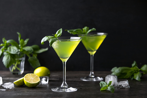 Basil Gimlet cocktail in a cocktail glasses standing on a dark wooden bar countertop surface ready to drink