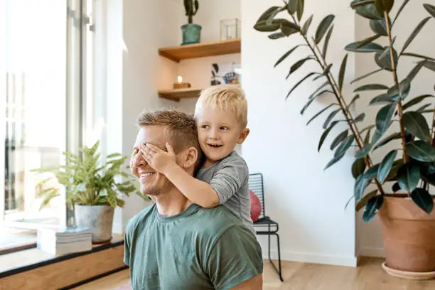 Smiling preschool boy covering eyes of father in living room. Man is enjoying weekend with blond son. They are spending leisure time together.
