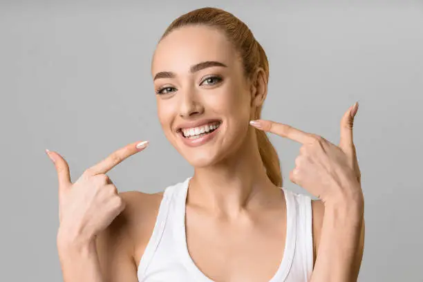 Orthodontic concept. Happy girl showing her beaming white teeth with two forefingers, grey background
