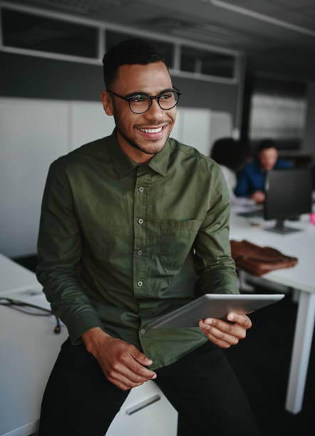 jeune homme d'affaires de sourire pensif retenant la tablette numérique regardant loin tout en s'asseyant à son lieu de travail dans le bureau - technology contemplation clothing formalwear photos et images de collection