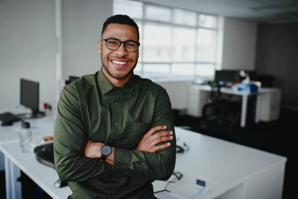Portrait of a happy confident young african american businessman standing with his arms crossed looking at camera Portrait of young and successful businessman in modern office expertise stock pictures, royalty-free photos & images