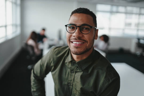 Portrait of charming successful young african american entrepreneur in shirt and eyeglasses looking at camera with smile in office Confident smiling young businessman against coworkers at background stubble male african ethnicity facial hair stock pictures, royalty-free photos & images