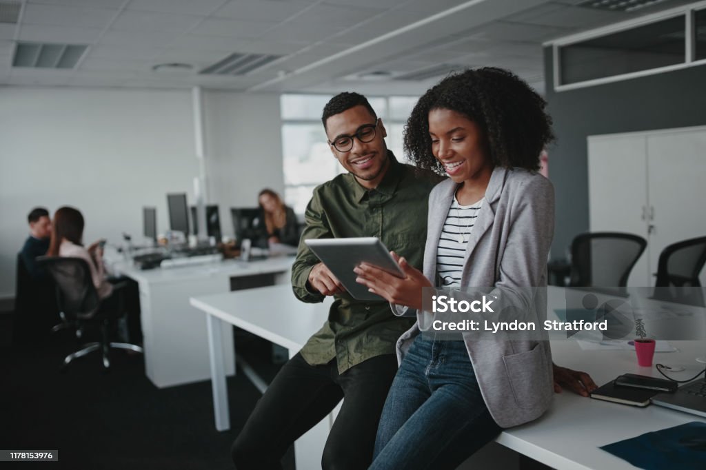 Successful two african american young businesspeople sitting on desk using digital tablet while colleague in background at office Successful african american business couple talking and using digital tablet Working Stock Photo