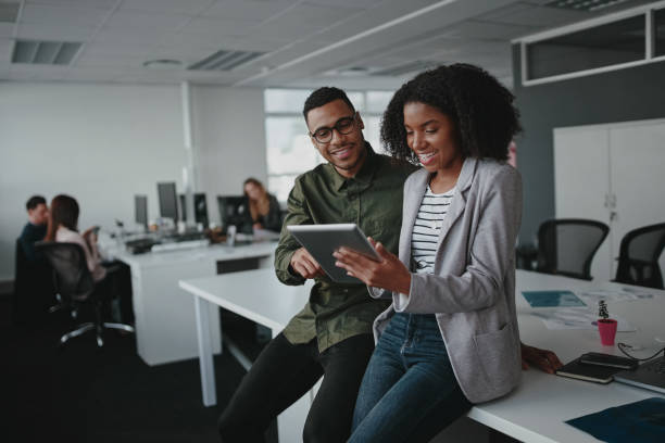 Successful two african american young businesspeople sitting on desk using digital tablet while colleague in background at office Successful african american business couple talking and using digital tablet coworker stock pictures, royalty-free photos & images