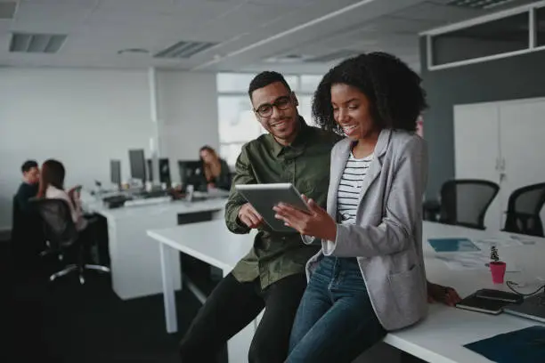 Photo of Successful two african american young businesspeople sitting on desk using digital tablet while colleague in background at office