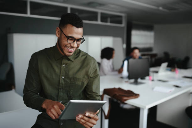 jeune homme d'affaires gai utilisant le sourire numérique de tablette tandis que ses collègues travaillant à l'arrière-plan - planning business person clothing well dressed photos et images de collection