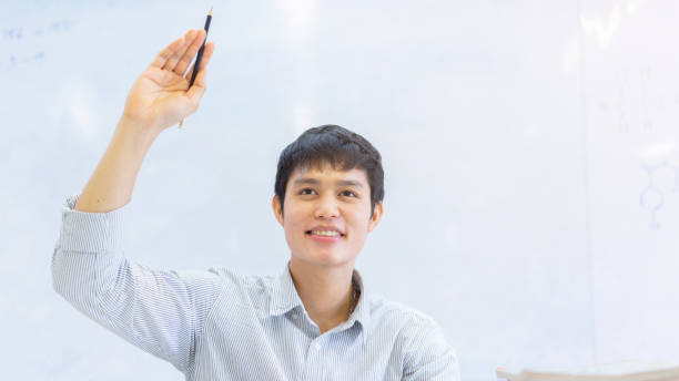 fermer le jeune homme asiatique d'étudiant d'université lèvent la main à demander l'enseignant au sujet du projet ou de l'examen dans la salle de classe pour l'éducation et le concept de personnes - school supplies education school equipment photos et images de collection