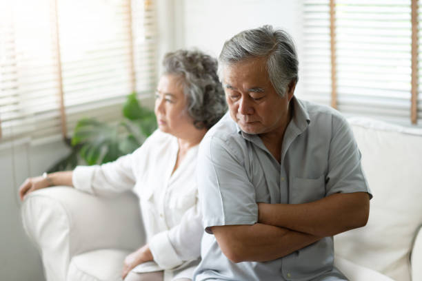 upset senior man sitting on sofa against the background of his wife. - arguing senior adult conflict couple imagens e fotografias de stock