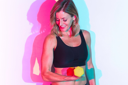 Waist up / one person of 20-29 years old adult beautiful caucasian female / young women standing / exercising in front of white background / multi-colored background / colored background wearing sports clothing / sports bra / shorts / running shorts who is smiling / happy / cheerful and being active with dumbbell / weightlifting / weight training / weights / cross training / body building / hand weight with shadow / gel effect lighting