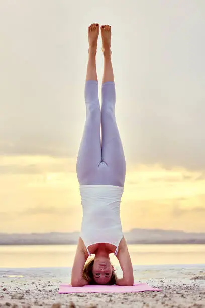 Woman in sportswear perform asana on nature near salt lake during picturesque sunset, yogi female doing Hatha yoga headstand or Sirsasana front full-length vertical view, balance of body mind concept
