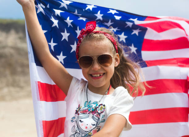 american girl with american flag. little smiling patriotic girl holding an american flag waving in the wind on the ocean beach. national 4 july. memorial day. independence day. flag day - child flag fourth of july little girls imagens e fotografias de stock