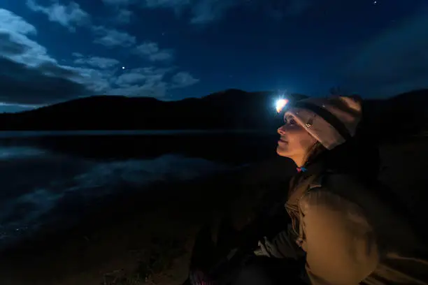 A young woman is looking at the clear sky at night in camping. You can see the reflection of the moon, the stars and the clouds on the lake. She is wearing a hat and a headlamp. The location is Mont-Tremblant, Quebec, Canada