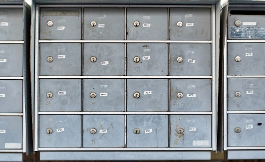 Apartment mailboxes lined up in a row. The numbered boxes are metal and appear very worn out with scratches, dirt and other markings.