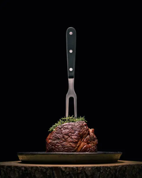 Photo of Sous-vide grilled beef steak with fork and herbs on dark background.