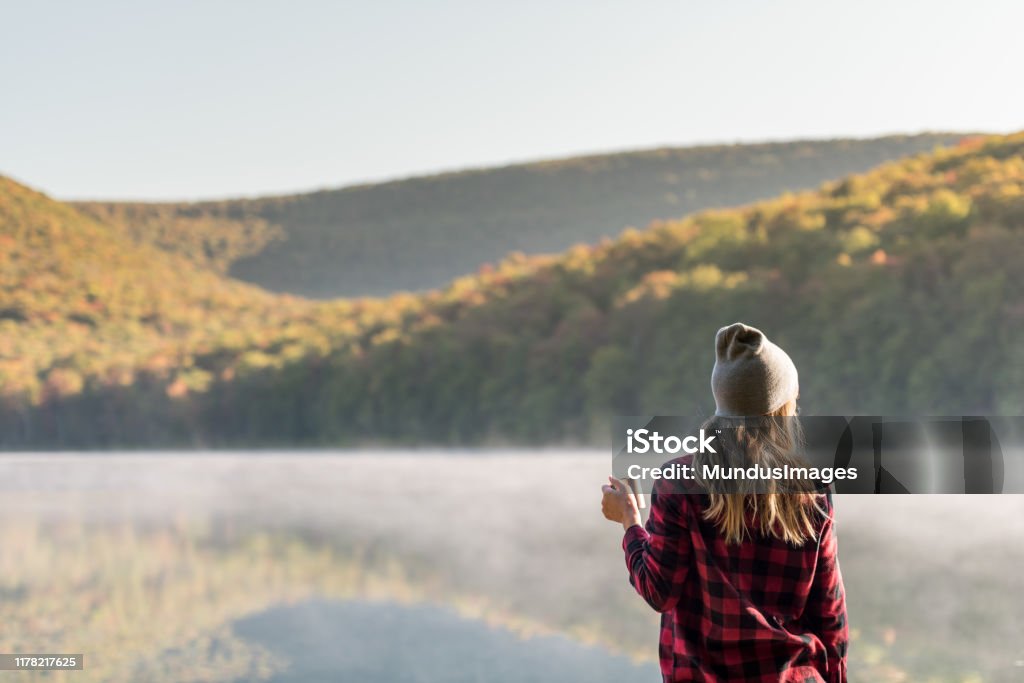 Young woman hiking with coffee A young woman is hiking and walking with coffee while camping at a lake in the morning. Autumn Stock Photo