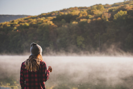 A young woman is hiking and walking with coffee while camping at a lake in the morning.