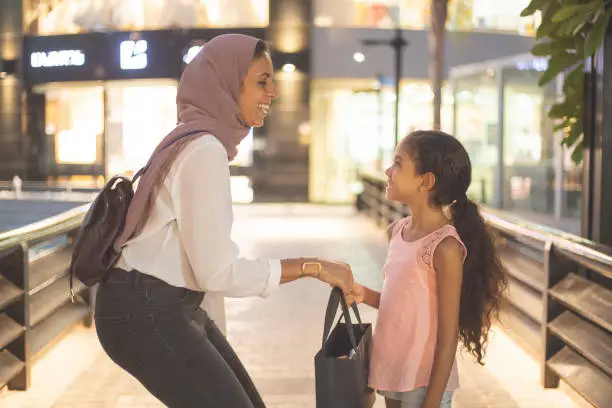 A Muslim mother and daughter are in a shopping mall. The mother is passing her purse to the girl.