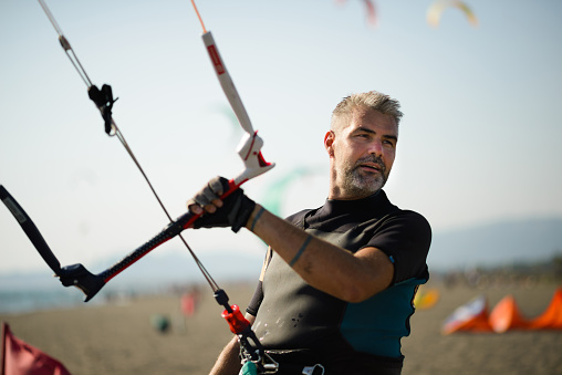 Mature adult gray-haired man is holding on to a bar and holding his kite in the air on a kitesurfing beach