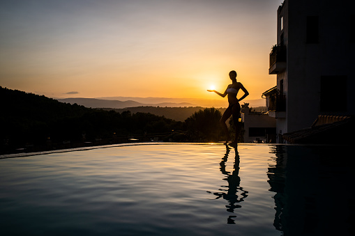 Silhouette and reflection of woman with ideal figure holding on a sun at infinity luxury pool at sunset