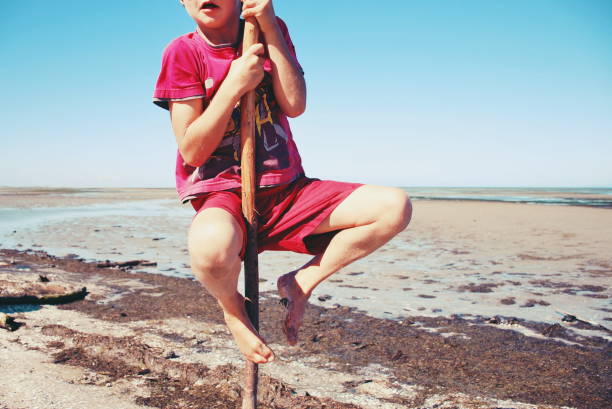 Horizontal photograph of a small child playing with beach toys on sand of a beach in England. The boy in a red vest and shorts is sitting in a small pit and digging sand with a play shovel. A wheelbarrow and bucket and other toys lie along on the sand. Copy space for text.