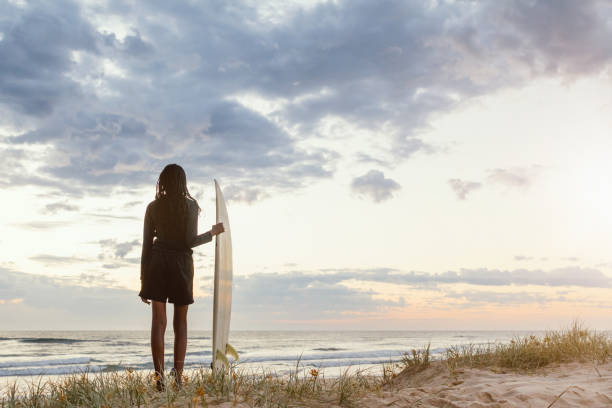 This photo captures the serene beauty of a tropical sunset over the ocean. The sky is vibrant shades of orange, pink and yellow, with dramatic clouds that create a unique atmosphere. Colorful surfboard on the beach sand.