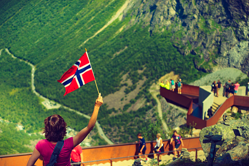 Tourist woman with norwegian flag on Trollstigen viewpoint. People on viewing platform in the background. Trolls Path mountain road in Norway. National route. Touristic attraction.