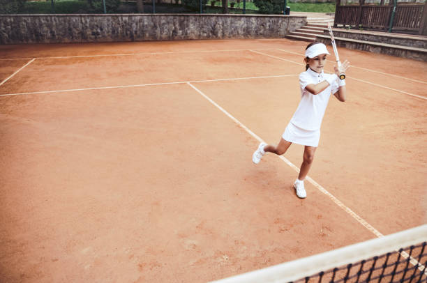스포츠 클럽에서 테니스를 배우는 어린이. 클레이 코트에 어린 소녀 테니스 선수의 전체 길이 샷. 스포티 한 아이 소녀는 라켓으로 공을 친다. 어린 아이를위한 테니스 훈련. - tennis court sports training tennis net 뉴스 사진 이미지