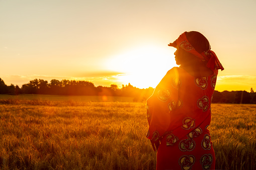 Mujer africana con ropa tradicional de pie, mirando, mano a los ojos, en el campo de cultivos de cebada o trigo al atardecer o al amanecer photo