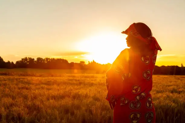 Photo of African woman in traditional clothes standing, looking, hand to eyes, in field of barley or wheat crops at sunset or sunrise