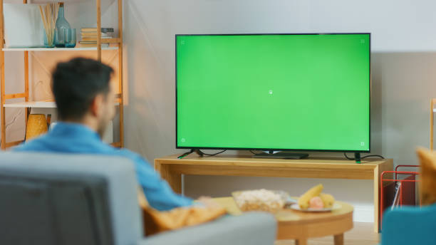 ragazzo felice seduto a casa nel soggiorno sulla sua sedia, guardando lo schermo verde della chiave chroma, rilassandosi dopo il lavoro. uomo in una stanza accogliente guardando partite sportive, notizie, programmi tv o film. - home movie foto e immagini stock