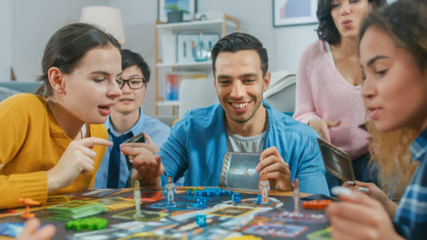 groupe diversifié de gars et de filles jouant dans un jeu de société stratégique unique avec des cartes et des dés. amis ayant des cartes de lecture de plaisir, plaisantant, faisant des mouvements et riant dans une salle de séjour confortable - meeple photos et images de collection