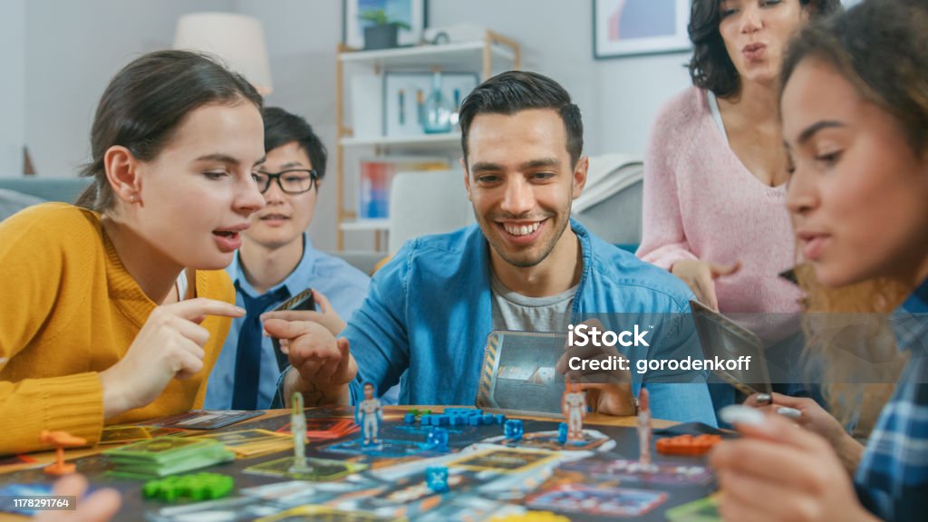 Diverse Group of Guys and Girls Playing in a Strategic Uniquely Designed Board Game with Cards and Dice. Friends Having Fun Reading Cards, Joking, Making Moves and Laughing in a Cozy Living Room Board Game Stock Photo