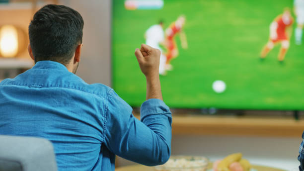 At Home Sports Fan Watches Important Soccer Match on TV, He Aggressively Clenches the Fist, Cheering for His Team. Cozy Living Room with Snacks and Drinks on the Table. At Home Sports Fan Watches Important Soccer Match on TV, He Aggressively Clenches the Fist, Cheering for His Team. Cozy Living Room with Snacks and Drinks on the Table. tv game stock pictures, royalty-free photos & images