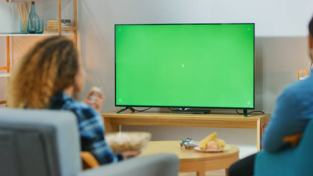 リビングルームで自宅に座って緑のクロマキースクリーンテレビを見て、ソファでリラックスして幸せなカップル。スポーツの試合、ニュース、ショーや映画を見てカップルルーム。 - テレビを見る ストックフォトと画像